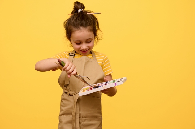 Smiling little girl artist dipping paintbrush into watercolor paints, isolated yellow background. Copy advertising space