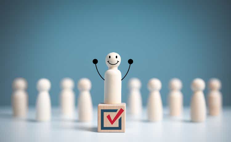 A wooden figure raising his hand stands on a wooden cube with different voting symbols on it.  Open mind or public hearing or election concept.  Volunteers, political parties, candidates, constituency voters.