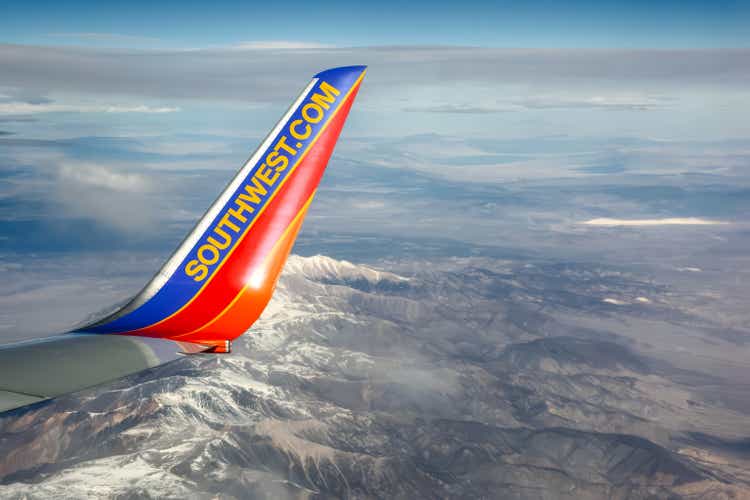 A wing tip of Southwest Airlines Boeing 737 flying over snow-capped mountains. The blue and orange colors contrast with snowy peaks and clear sky. The image captures Southwest