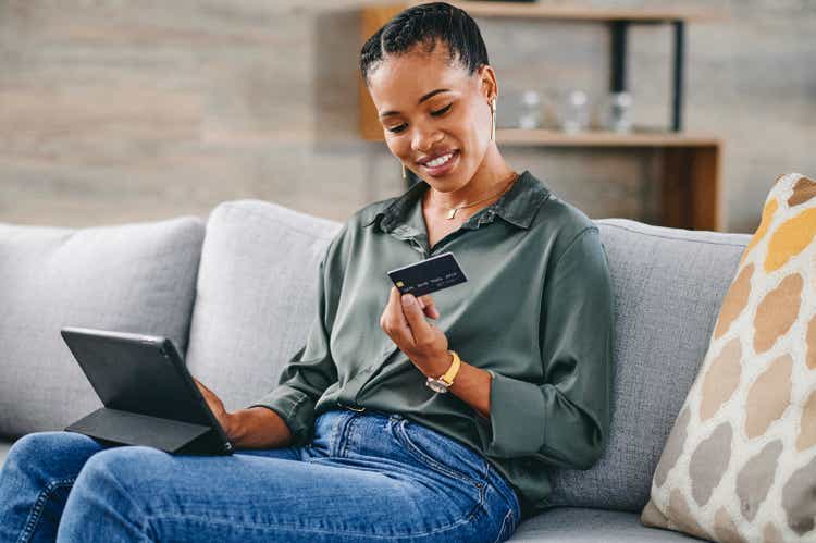 Happy woman shopping online with digital tablet, looking at credit card on sofa