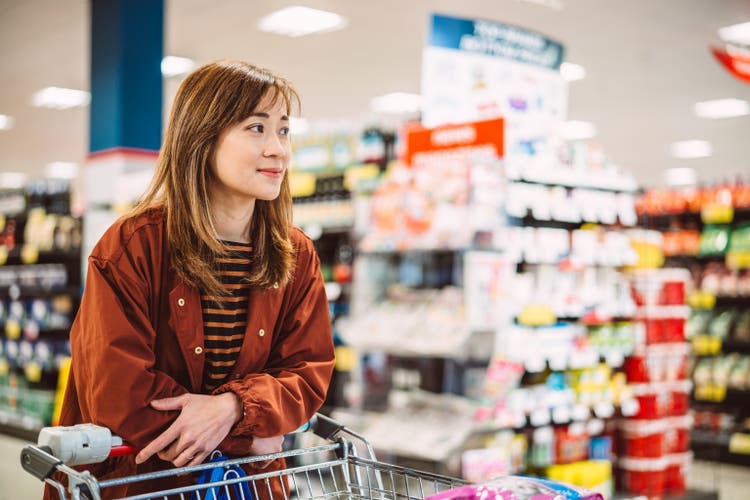 Young pretty Asian woman waiting in line for checkout while shopping for groceries in supermarket