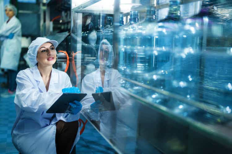 scientist worker checking the quality of water bottles on the machine conveyor line at the industrial factory. Female worker recording data at the beverages manufacturing line production.