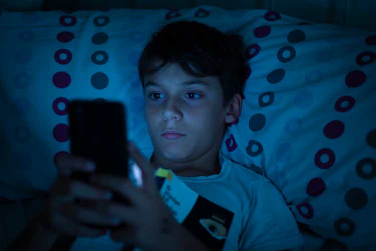 Child in bed at night with a book and a smartphone in the dark. Concept of problems with mobile addiction at young ages. Child with sleeping problems and insomnia due to the use of electronic devices