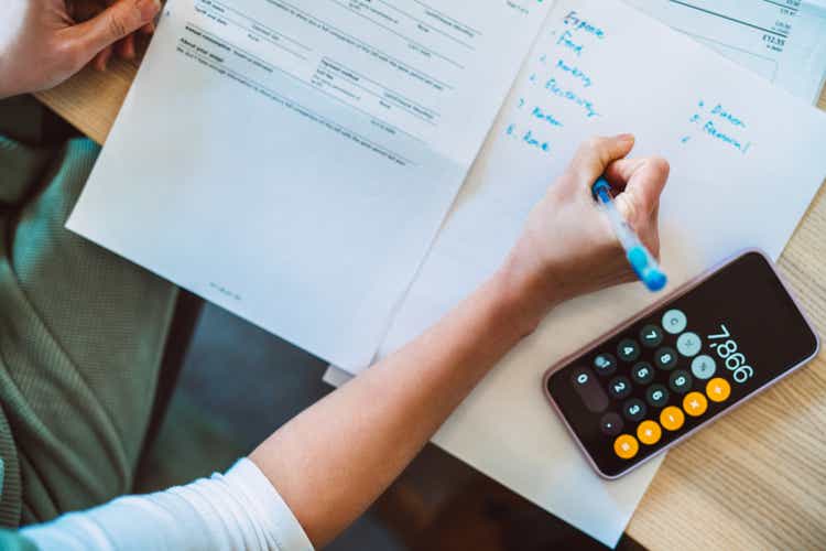 Overhead view of young Asian women managing home finance using smartphone. She is working with household utility bill and calculating expenses at home.