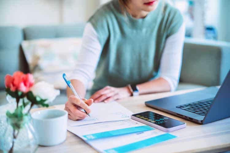 Young Asian women managing home finance using laptop & smartphone. She is working with household utility bill and calculating expenses at home.