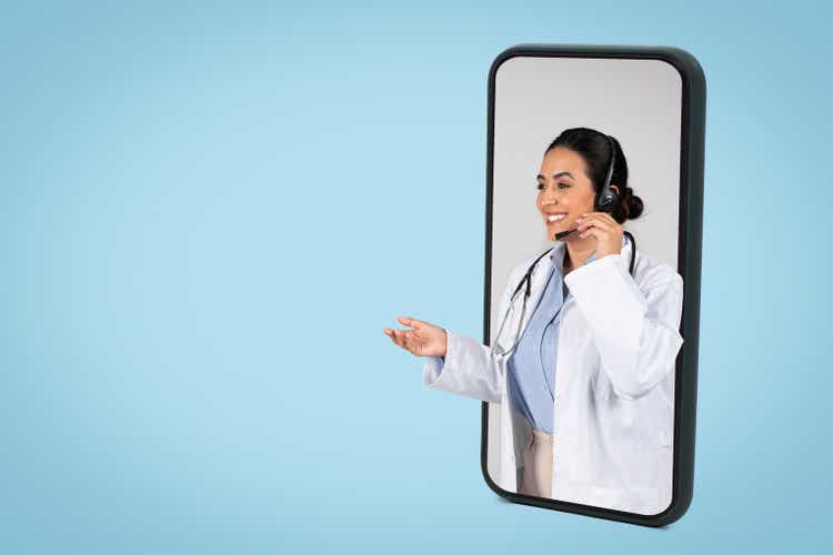 Happy latin woman doctor with headset in big cellphone screen having online appointment, blue background, copy space