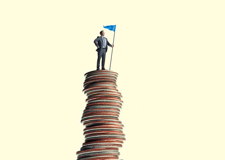 Businessman Stands With Flag On Top Of Tall Stack Of Coins