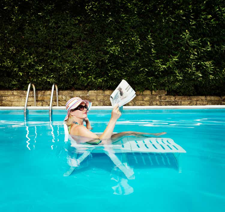 Woman in sun hat relaxing on sun lounger in pool reading newspaper