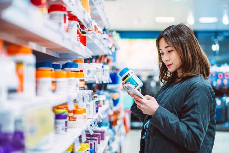 Beautiful Asian woman searching information about the product & technology using smart phone while shopping for multi vitamin & health supplements in supermarket. Heathy lifestyle concept.