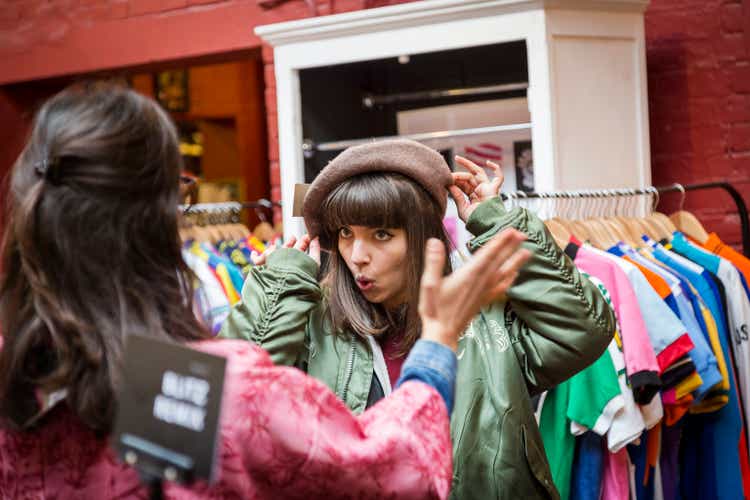 Woman tries on hat in shop, while friend looks on.