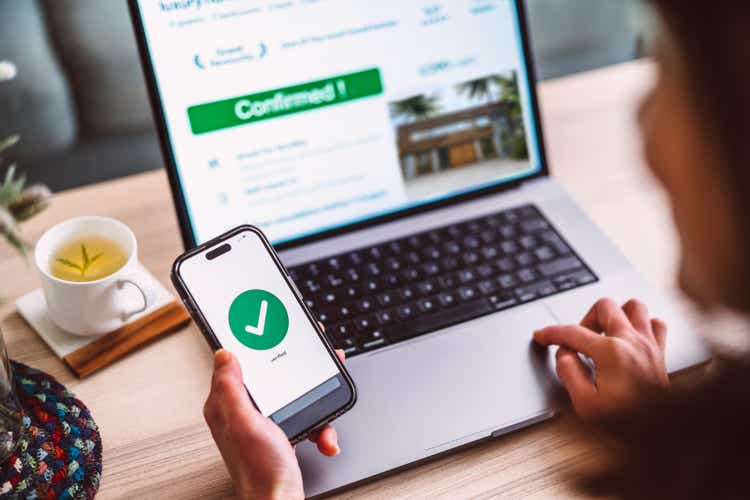 Cheerful Asian woman booking accommodation on travel app and paying through mobile app on smart phone at home