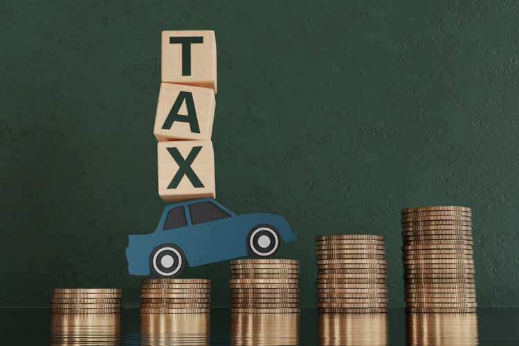 Wooden blocks forming the word TAX on a green paper cut car running on stacks of gold coins on green background. Illustration of the concept of government vehicle tax and related expenses