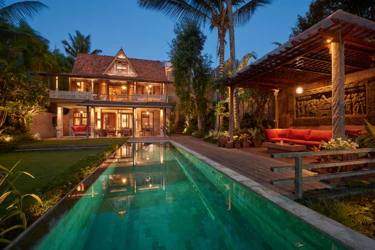 Traditional wooden house at dusk with Pergola sitting area in foreground next to pool, illuminated at night