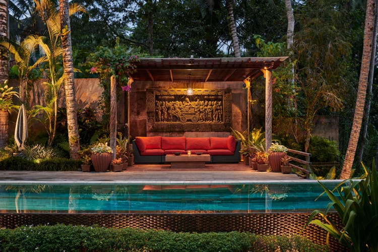 Pergola with sitting area next to pool at luxury villa, situated in garden with tropical plants at dusk
