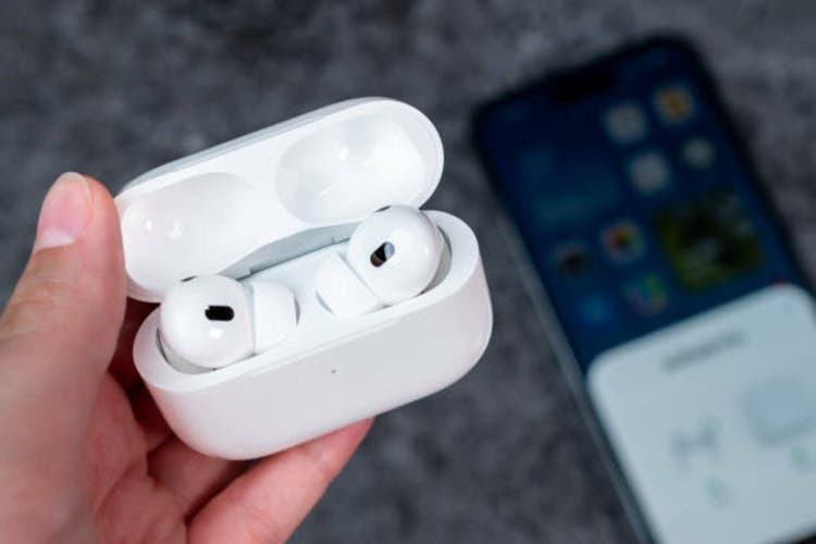 In woman hand there is open case with headphones - AirPods Pro 2 generation. On background out of focus smartphone witch are connected to Air Pods.