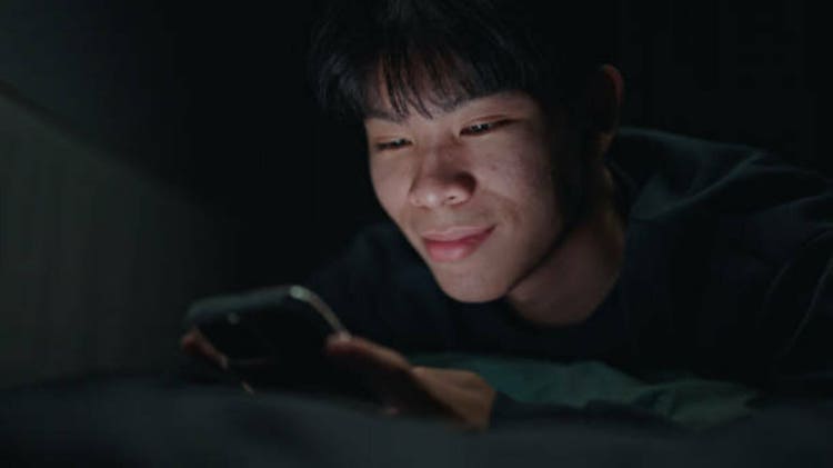 Teenage boy relaxing with smartphone in bedroom at night.