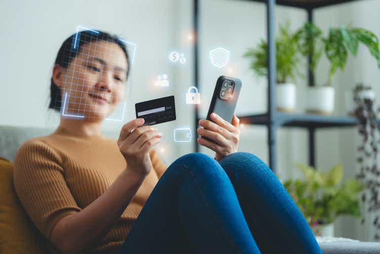 Asian woman holding credit card and scanning face id by smart phone for shopping online sitting on sofa in living room at home.