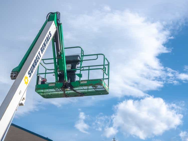 A Sunbelt Rentals aerial lift against a clear blue sky. The green and white lift platform is raised high, indicating it"s ready for use in construction.