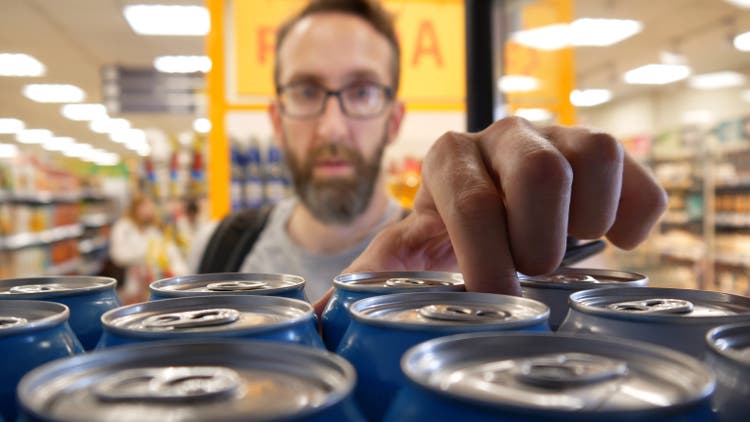 Lähikuva monista alumiinitölkeistä olutta kaupan jääkaapissa ja miespuolinen ostaja ottamassa yhden
