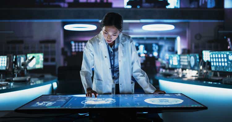 Female Asian neuroscinetist uses interactive touchscreen table with MRI scans on display in modern biotechnology research center. Doctor develops innovative devices for people with Alzheimer's