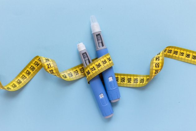 Two injectors dosing pens for subcutaneous injection of antidiabetic medication or anti-obesity medication hovering over a blue background. Yellow measuring tape around the injectors.