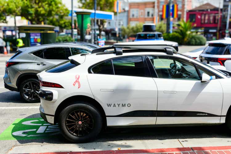 A white Waymo self-driving Jaguar I-PACE, adorned with a pink breast cancer awareness ribbon, with other vehicles in the background