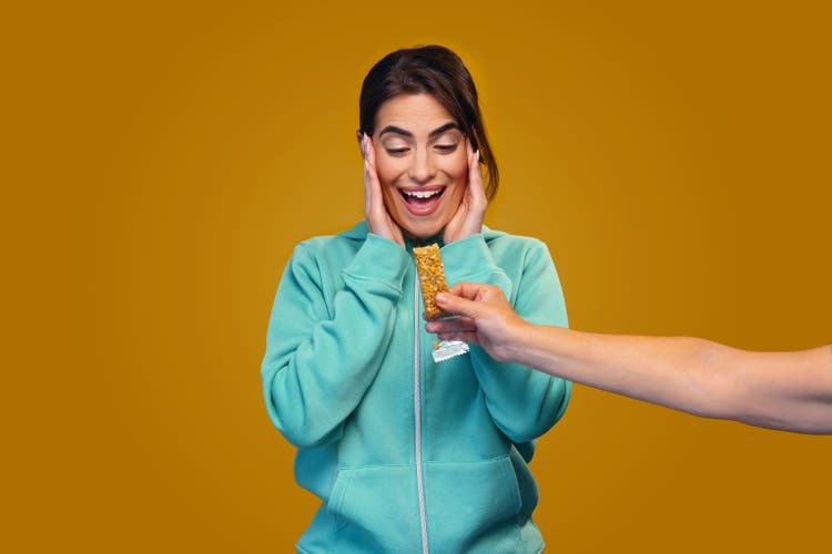 Cheerful smiling woman with blue sweater holding energy bar while posing in front of camera on isolated orange