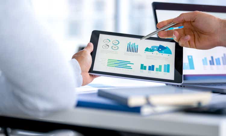 Two business men meeting and looking at financial data, charts and graphs on a digital tablet and a laptop computer.