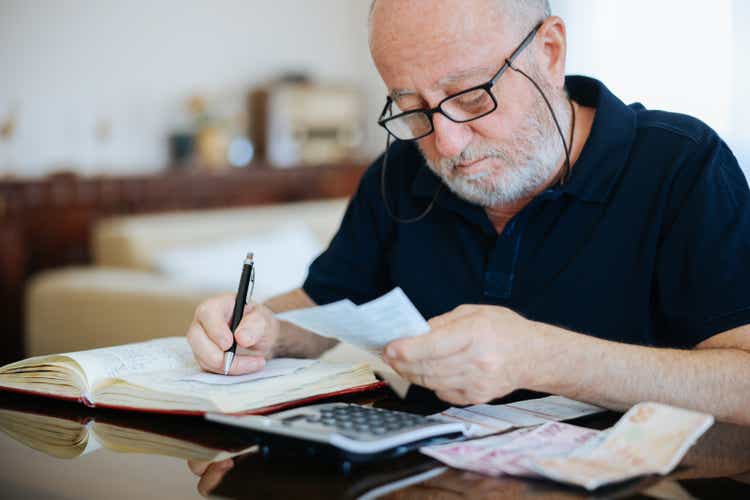 Anxious senior man managing finances, holding cash and using calculator at home