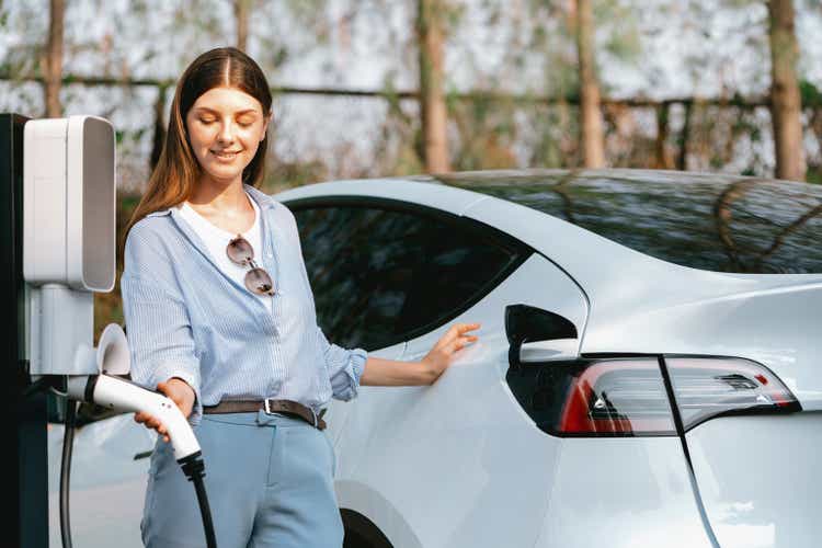 Young woman recharging battery for electric car in autumn. Exalt