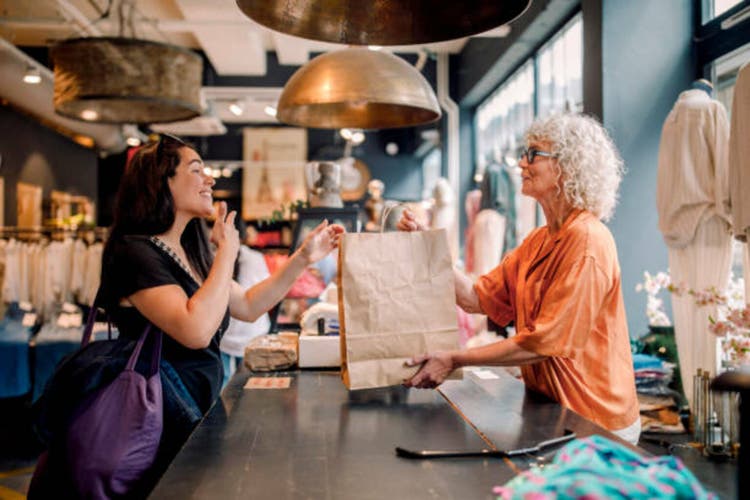 Senior female owner giving bag to customer at checkout in clothing store