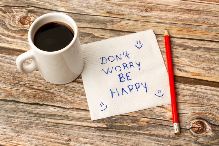 A mug with coffee with a paper with dont worry be happy phrase and a pencil on a wooden table
