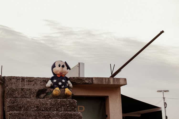 A Mickey Mouse stuffed doll stands at the stairs of an old house with grey sky at the background.