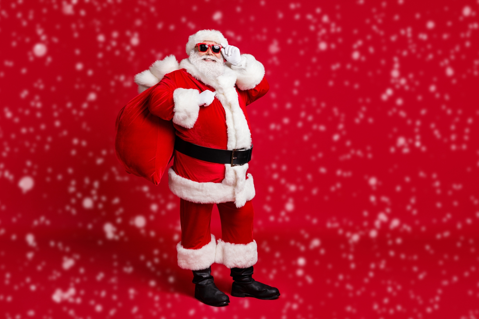 Full length photo of retired old man grey beard hold glasses bear sack presents good children wear santa costume gloves coat belt sunglass headwear black boots isolated red color background