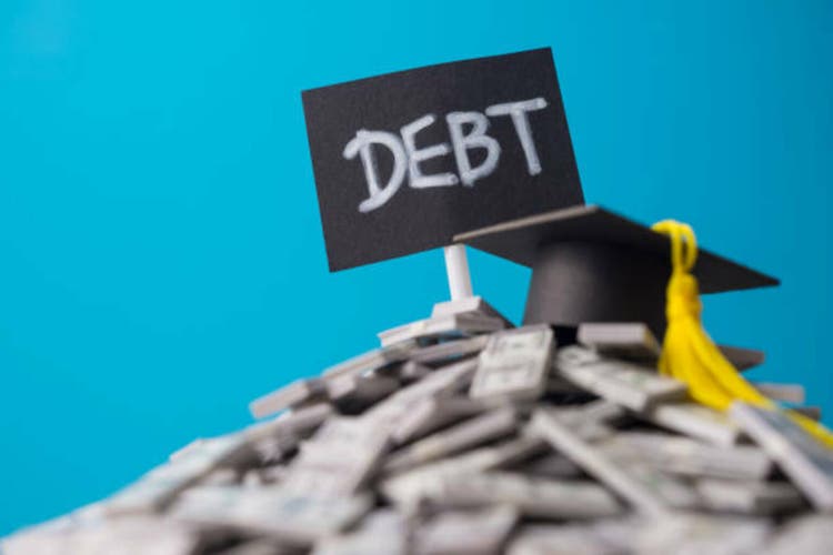 Mortarboard on heap US dollar banknotes and DEBT label with blue background.