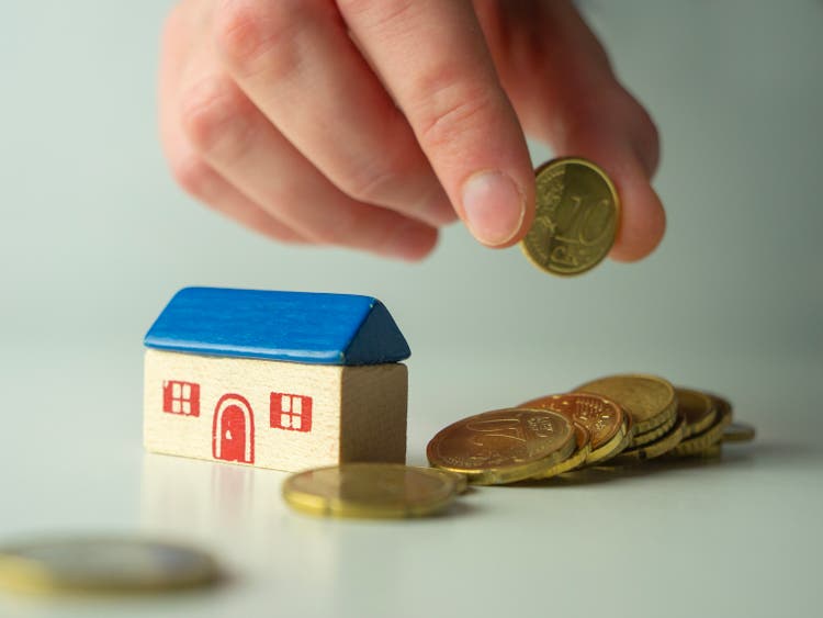 hand puts a coin on the pile of coins next to the little house concept of value and expense for property real estate
