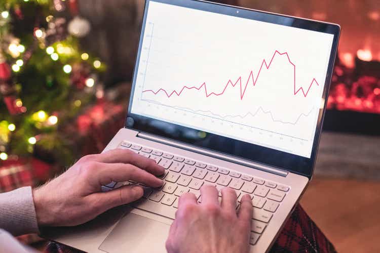 Man freelancer in santa claus hat working on laptop with graphs and charts on screen sitting near christmas tree and fireplace.