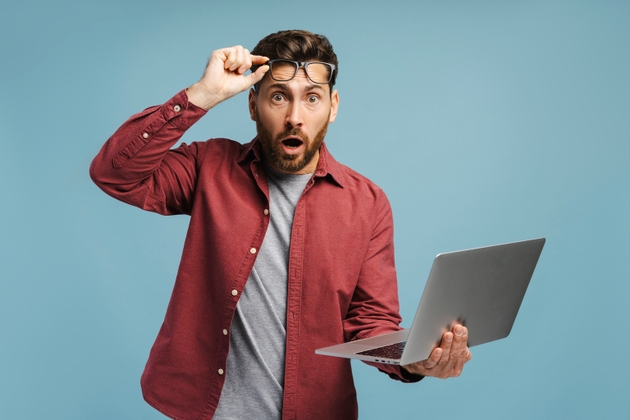 Shocked bearded Latin man wearing stylish eyeglasses, holding laptop, using computer with open mouth
