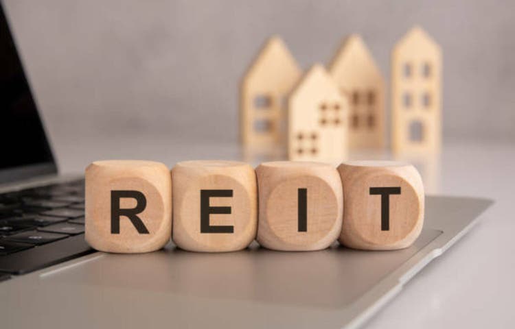 wooden cubes spelling "REIT" on a laptop, with wooden house models in the background, symbolizing real estate investment