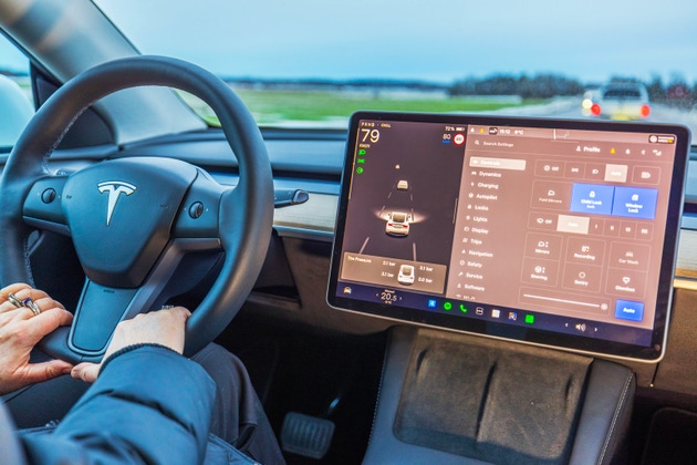 Close up view of Tesla Model Y driver holding steering wheel while using autopilot and control settings on touchscreen.