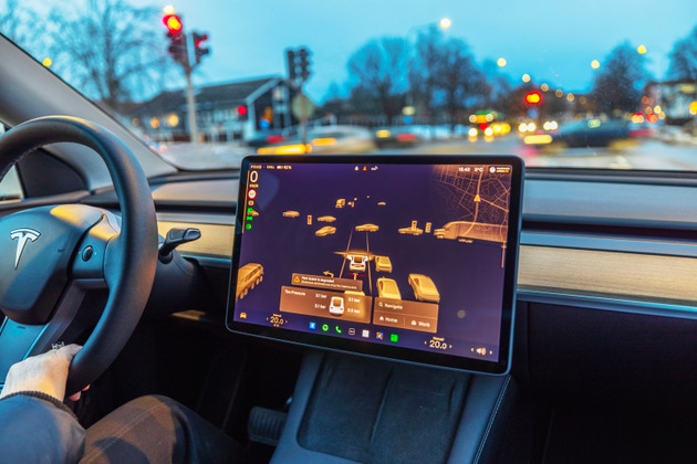 Close-up view of Tesla Model 3 interior with autopilot visualization on touchscreen while driver holds steering wheel.
