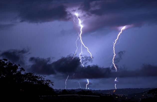 A dramatic lightning bolt illuminates the night sky above a suburban neighborhood, casting a purple glow over the horizon