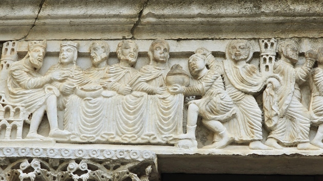 Nahaufnahme eines Steinreliefs, das sitzende Figuren an einem Tisch zeigt, mit einer knienden Figur, die ein rundes Gefäß anbietet, möglicherweise Teil einer religiösen oder historischen Erzählung.