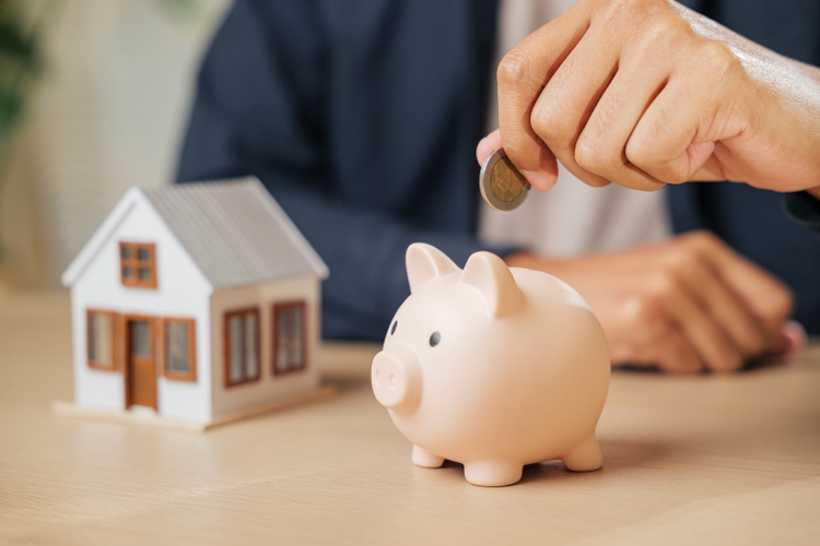 A businessman hand places a coin into a piggy bank beside a house model, symbolizing property savings, financial planning, and investment strategies for home buying