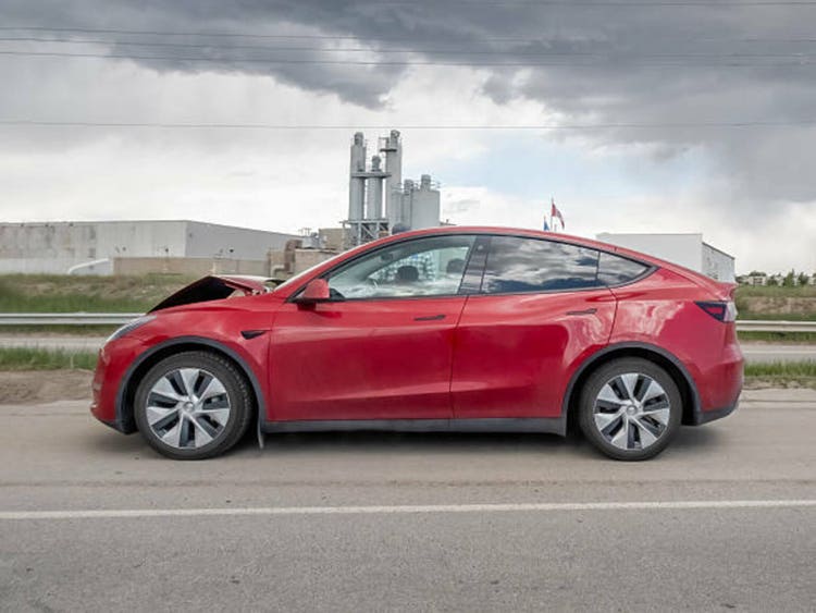 A striking red Tesla Model Y, seemingly involved in an accident, is parked roadside with its hood open, under a dramatic, cloudy sky and industrial backdrop.