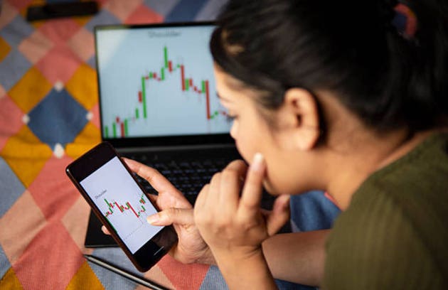 Gen Z young woman scrolling through investment trading data on smartphone and laptop.