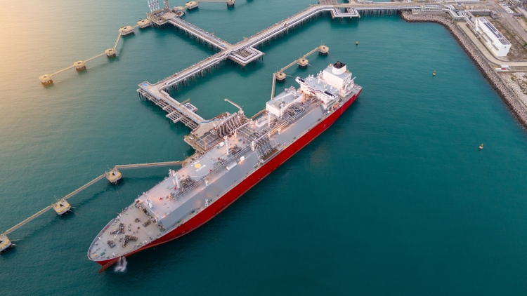 Aerial view of an LNG tanker docked at an offshore terminal with multiple pipelines, storage tanks, and an industrial complex. Captures global energy logistics