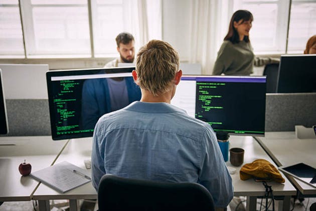 Rear view of male programmer coding on computers while sitting at desk in office