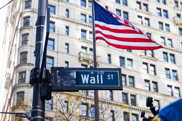 A classic street sign for Wall St stands prominently in a bustling urban setting, with the American flag waving above