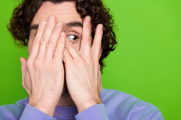 Curly-haired man in purple sweater covering face with hands on green background displaying amusement and curiosity